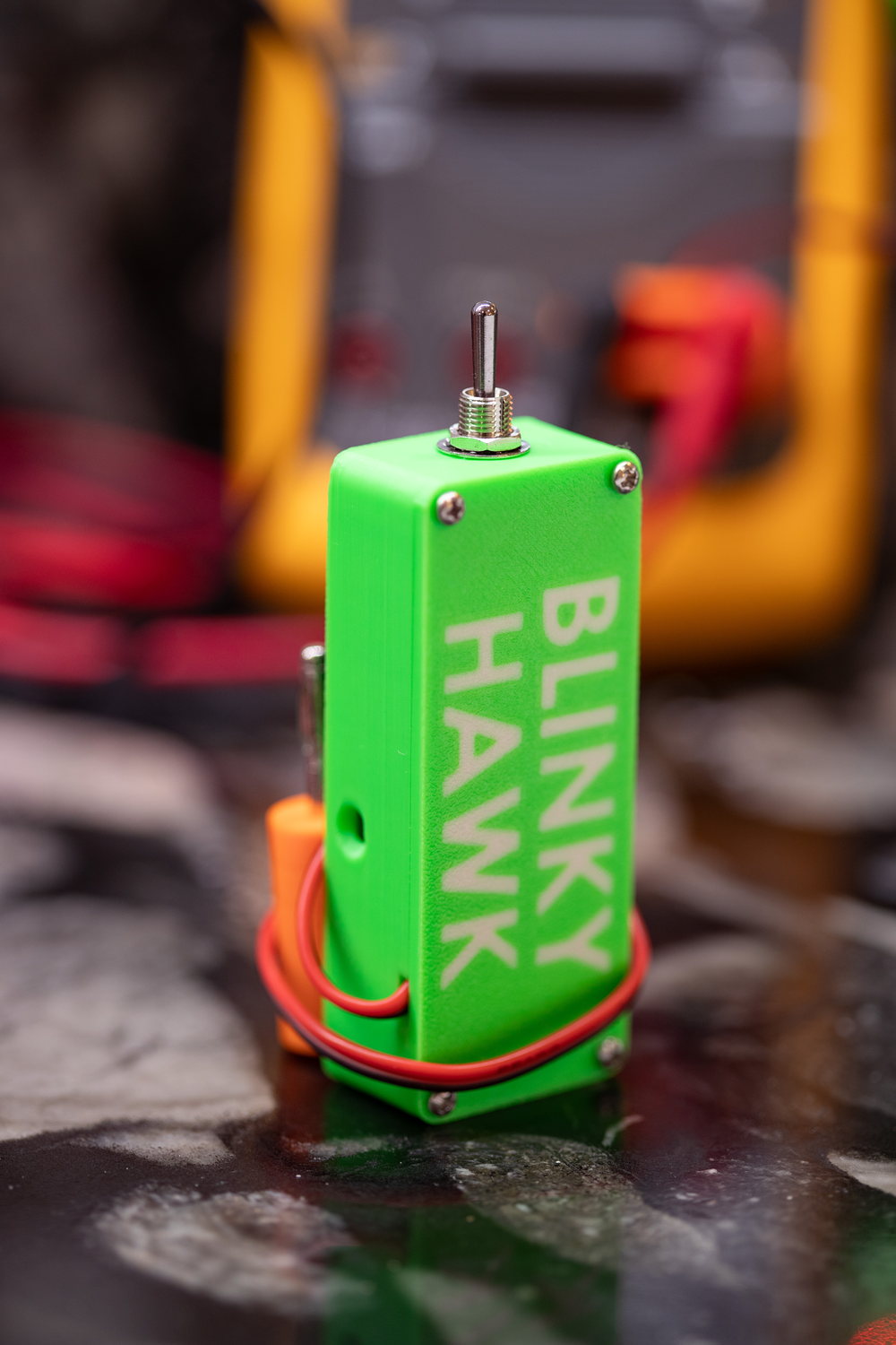 A Blinky Hawk standing on end on a countertop, the BLINKY HAWK name moulded into its green housing, with the metal toggle switch on top and an orange stacking banana plug and red lead at its base.