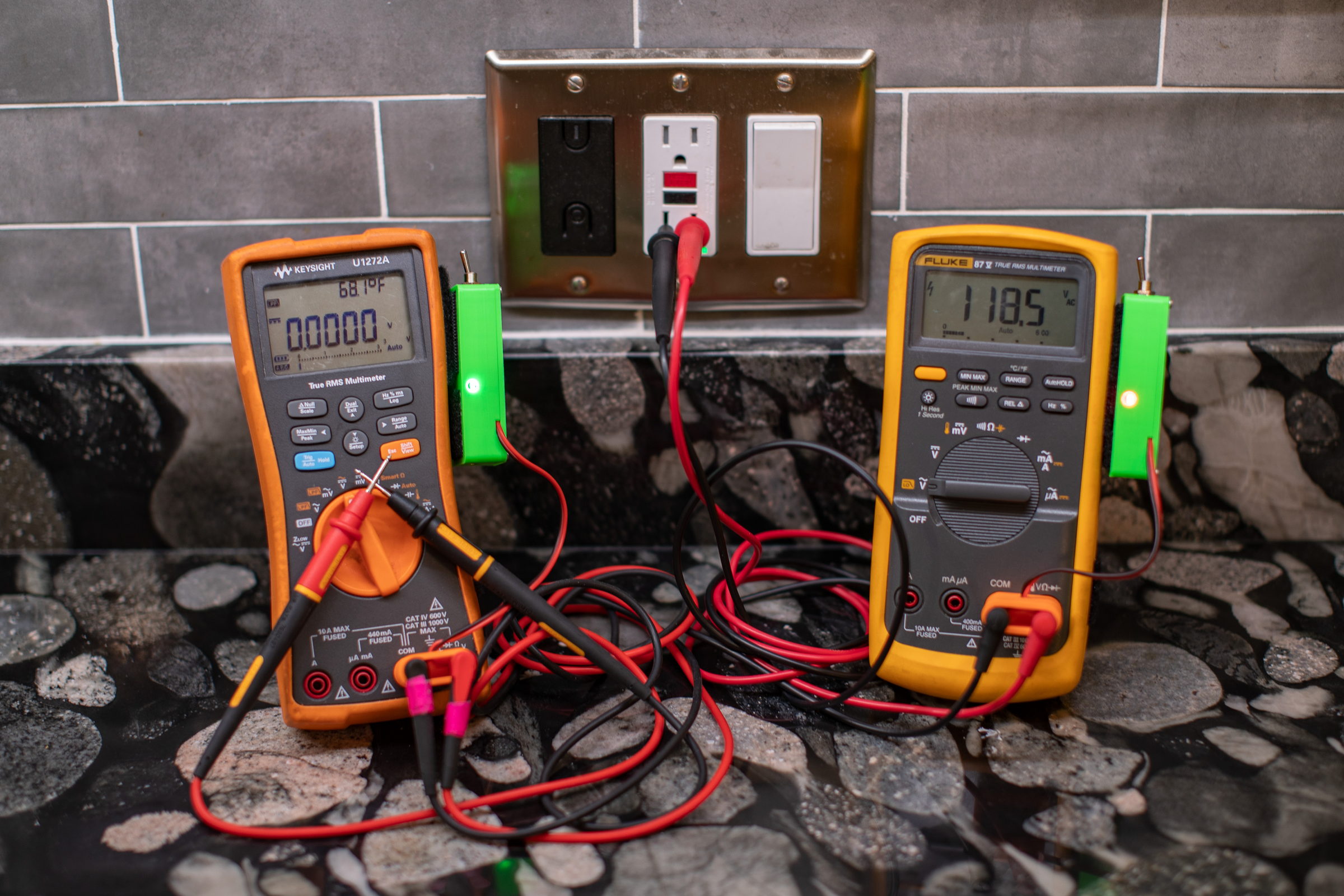 Two multimeters side by side on a countertop, each with a green Blinky Hawk attached. The left reads 0.0000 V with its detector lit green because its probes are touching; the right reads 118.5 V AC at a wall outlet with its detector lit orange.