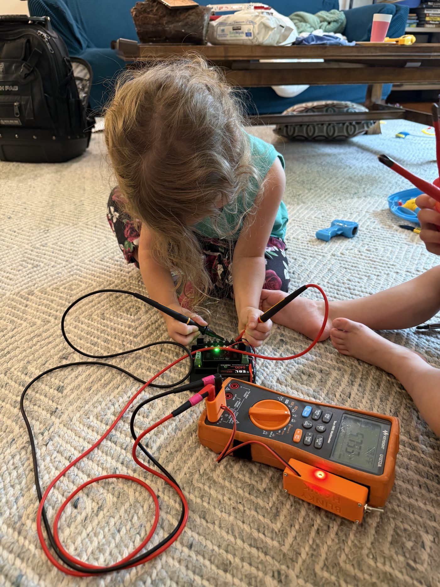 A small child kneeling on a rug, holding both multimeter probes onto a battery holder. The attached Blinky Hawk is lit orange to show voltage is present.