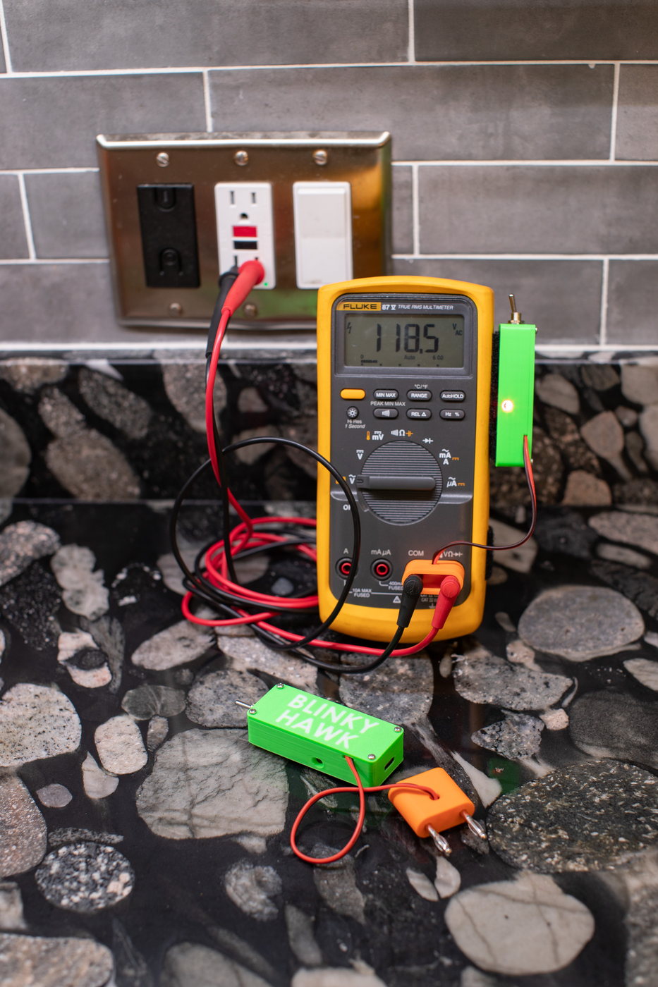 A Blinky Hawk unit lying on a countertop, the BLINKY HAWK name moulded into its green housing, with its stacking banana pins at one end, the buzzer port, the USB-C charging slot, a short red lead and a loose orange stacking banana plug beside it. Behind it a Fluke 87V reads 118.5 V AC at a wall outlet with a second unit attached, lit orange.