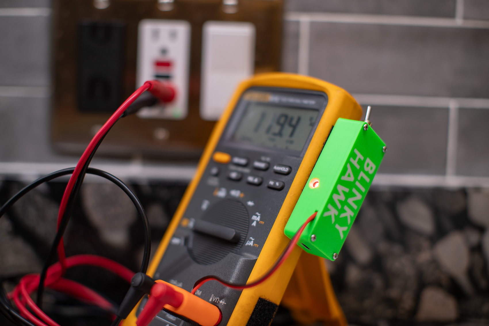 Close view of a Blinky Hawk riding on the upper edge of a yellow Fluke 87V. The meter reads 119.4 V AC and the small round indicator on the detector is lit orange.